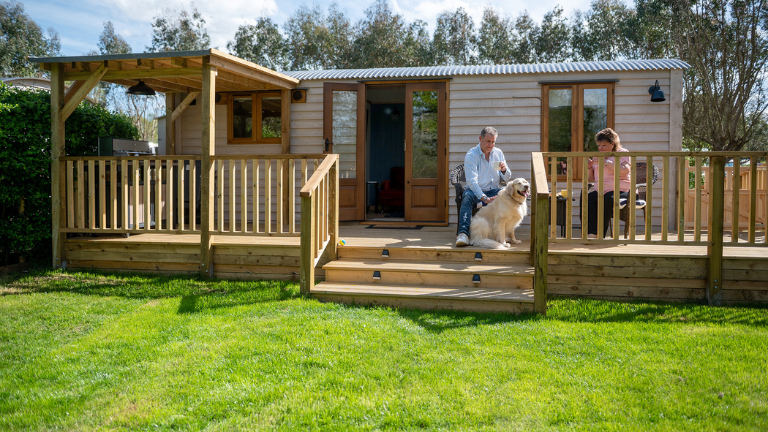 Two people and a dog sit on the decking area of an ensuite shepherd hut